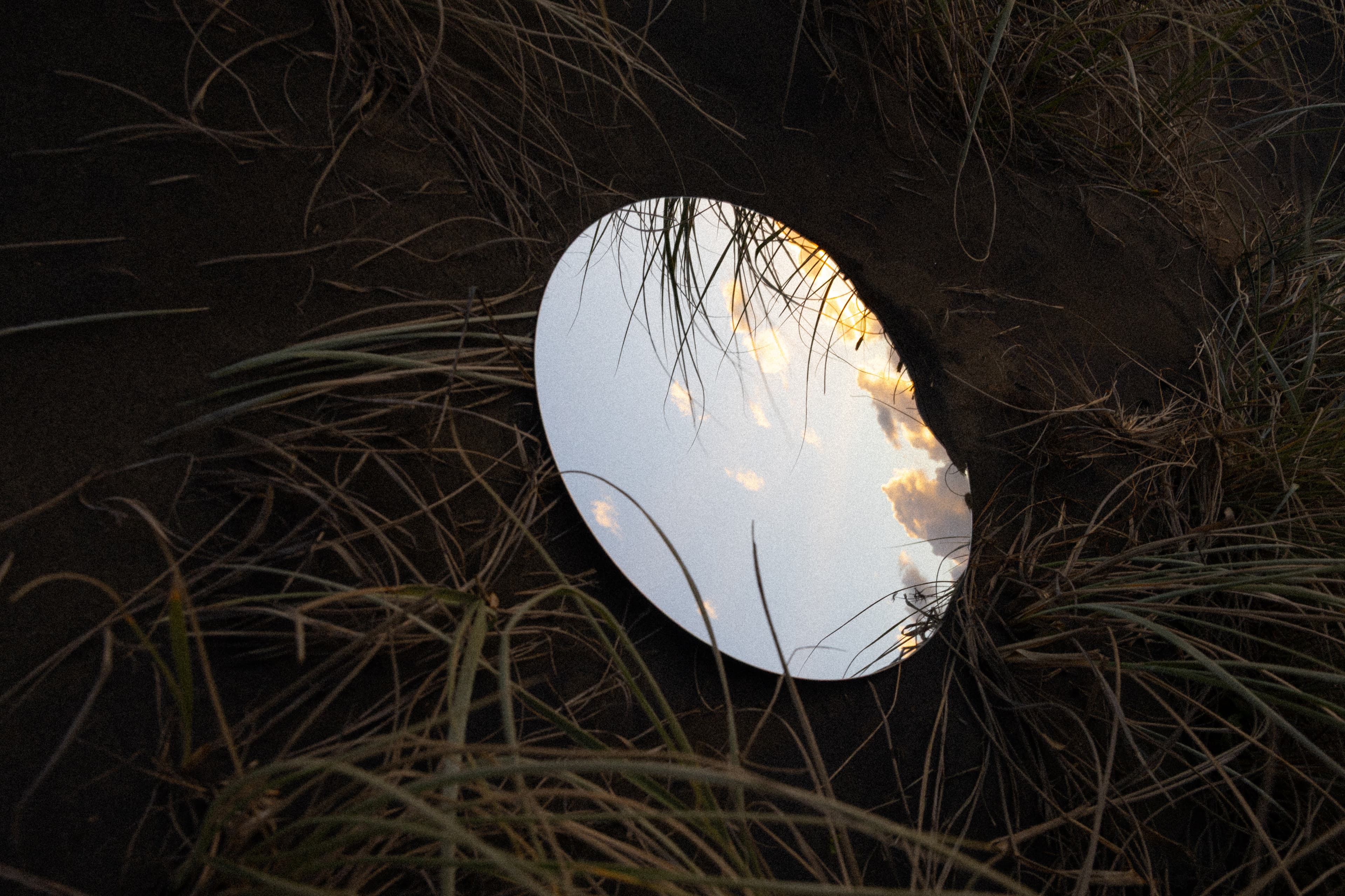 Circle shaped mirror sitting in sand dune grass reflecting the sky