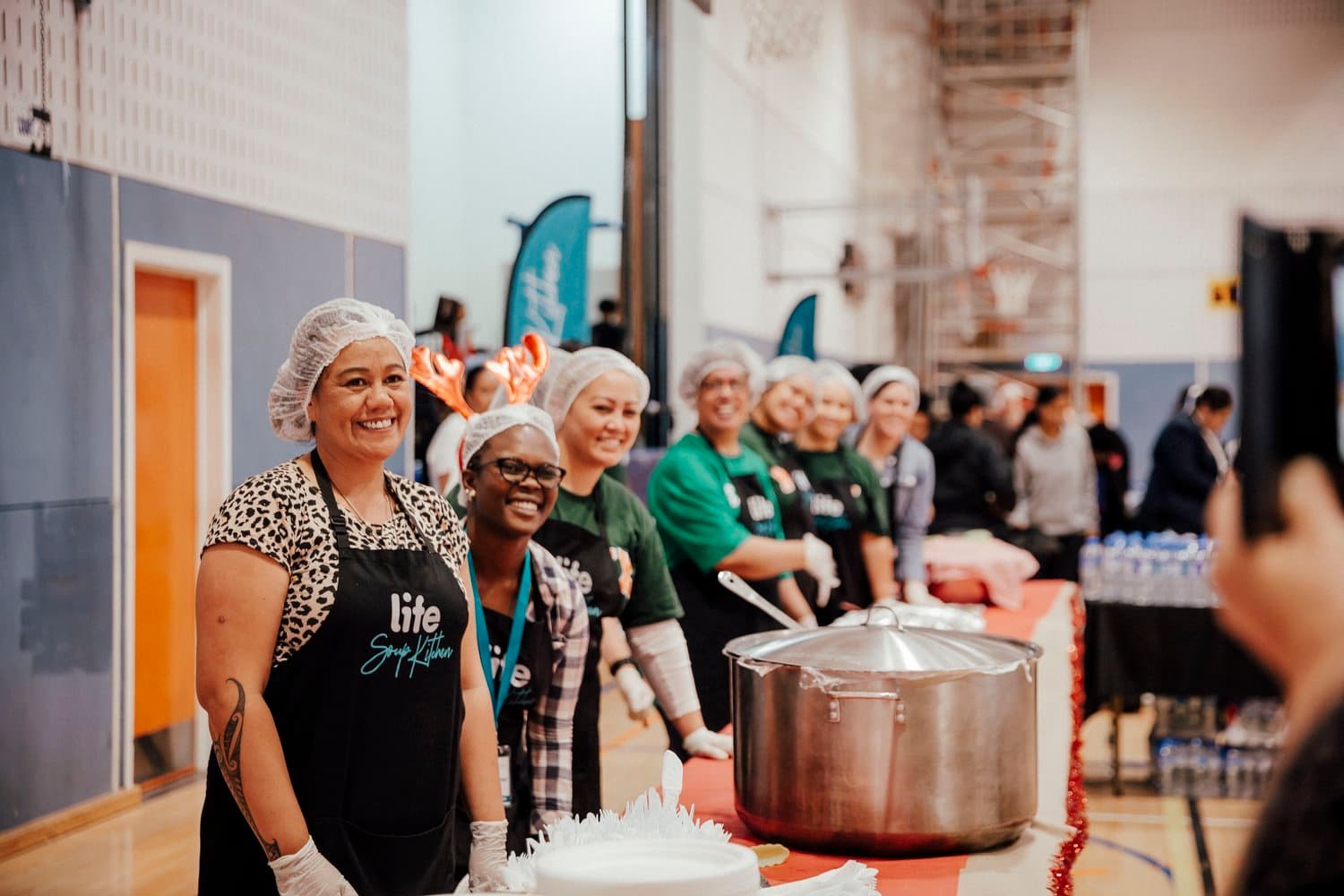 Team of people standing behind tables with food at LIFE Community Kitchen ready to serve, all wearing black aprons and hair nets