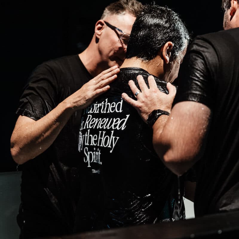 Back of person who has been baptised with the hands of the baptisers on his back, and t-shirt showing Titus 3:5 scripture on the back of the t-shirt