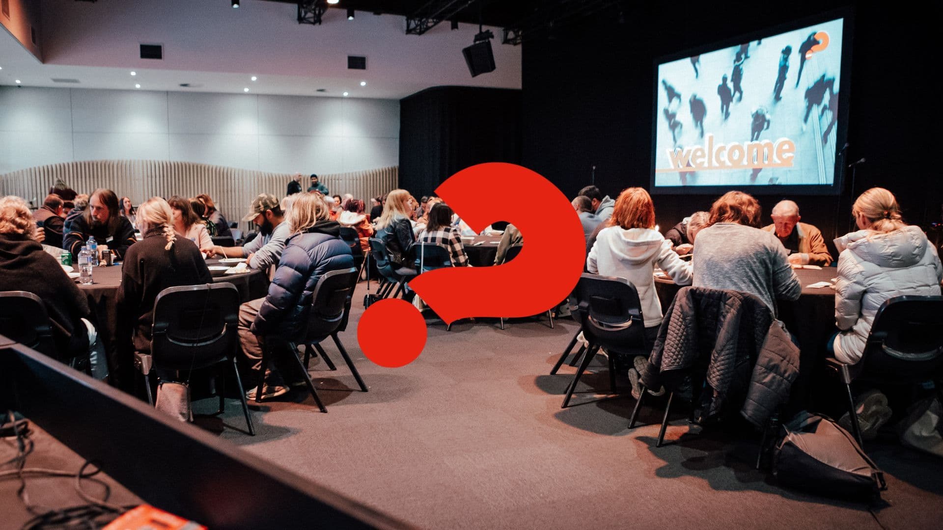 People sitting at round tables in the Chapel discussing the Alpha Course content, and the red Alpha Course brand question mark over the centre of the image