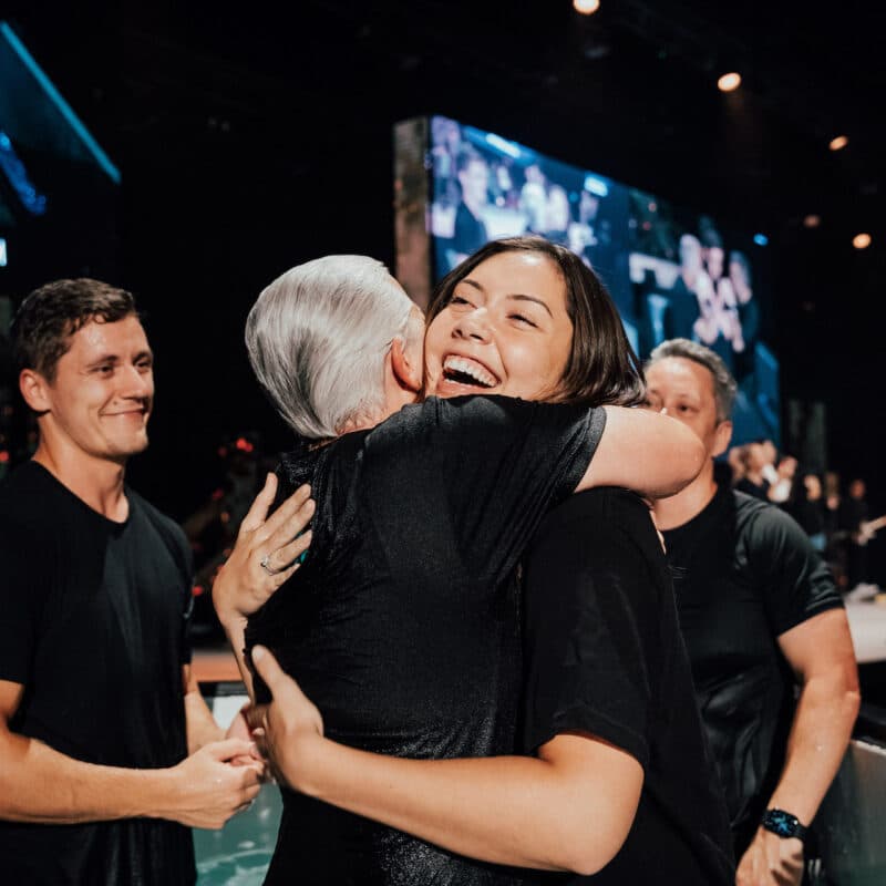 Two women hugging and smiling after one has been baptised, with the baptisers standing behind in the baptism pool with smiles