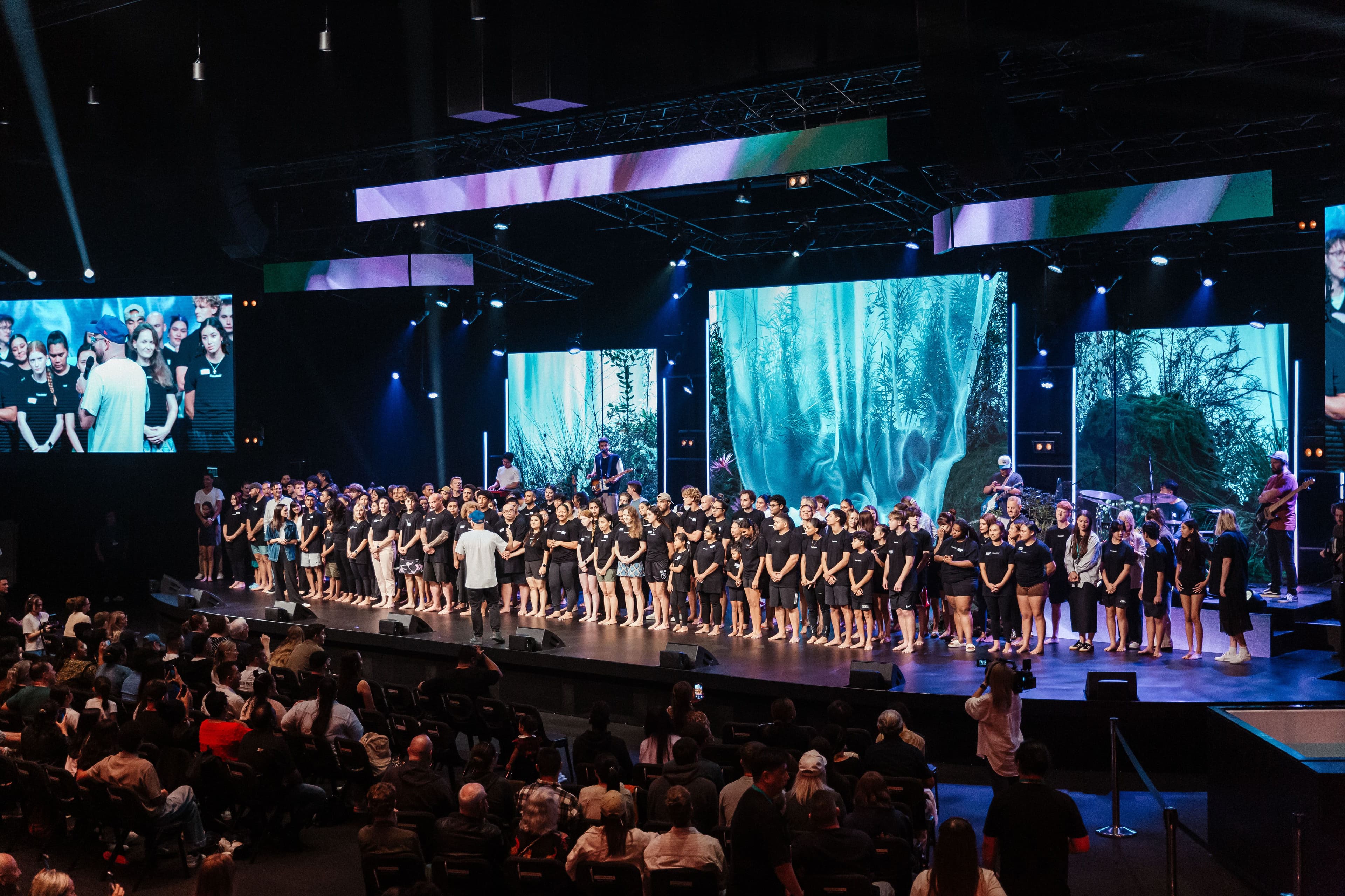 Everyone who is getting baptised standing in matching baptism t-shirts on the stage getting prayed for by the pastoral team before their baptism