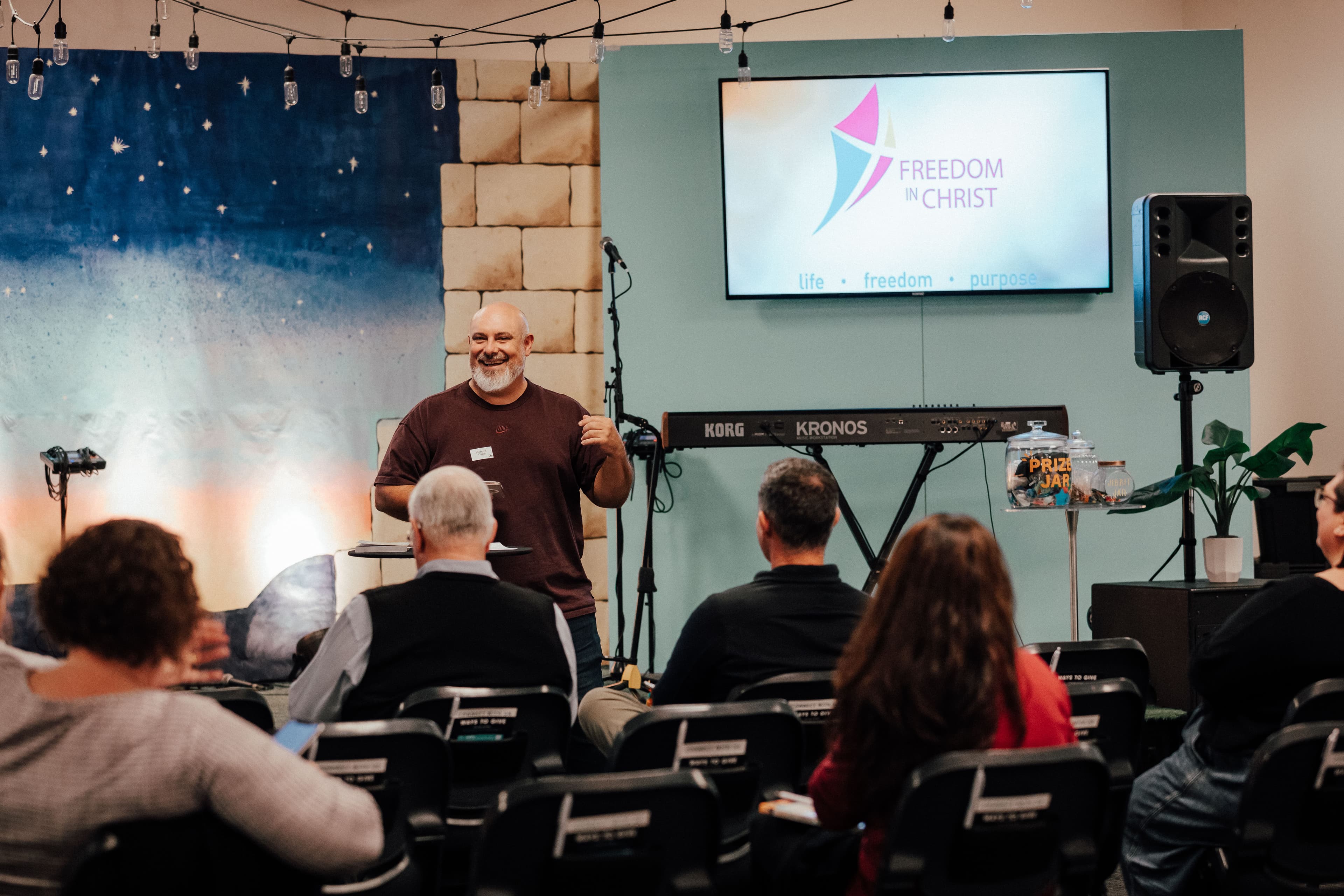 Facilitator of the Freedom Course talking to the attendees in the kids auditorium at LIFE North in Albany with a TV showing the Freedom content in the background