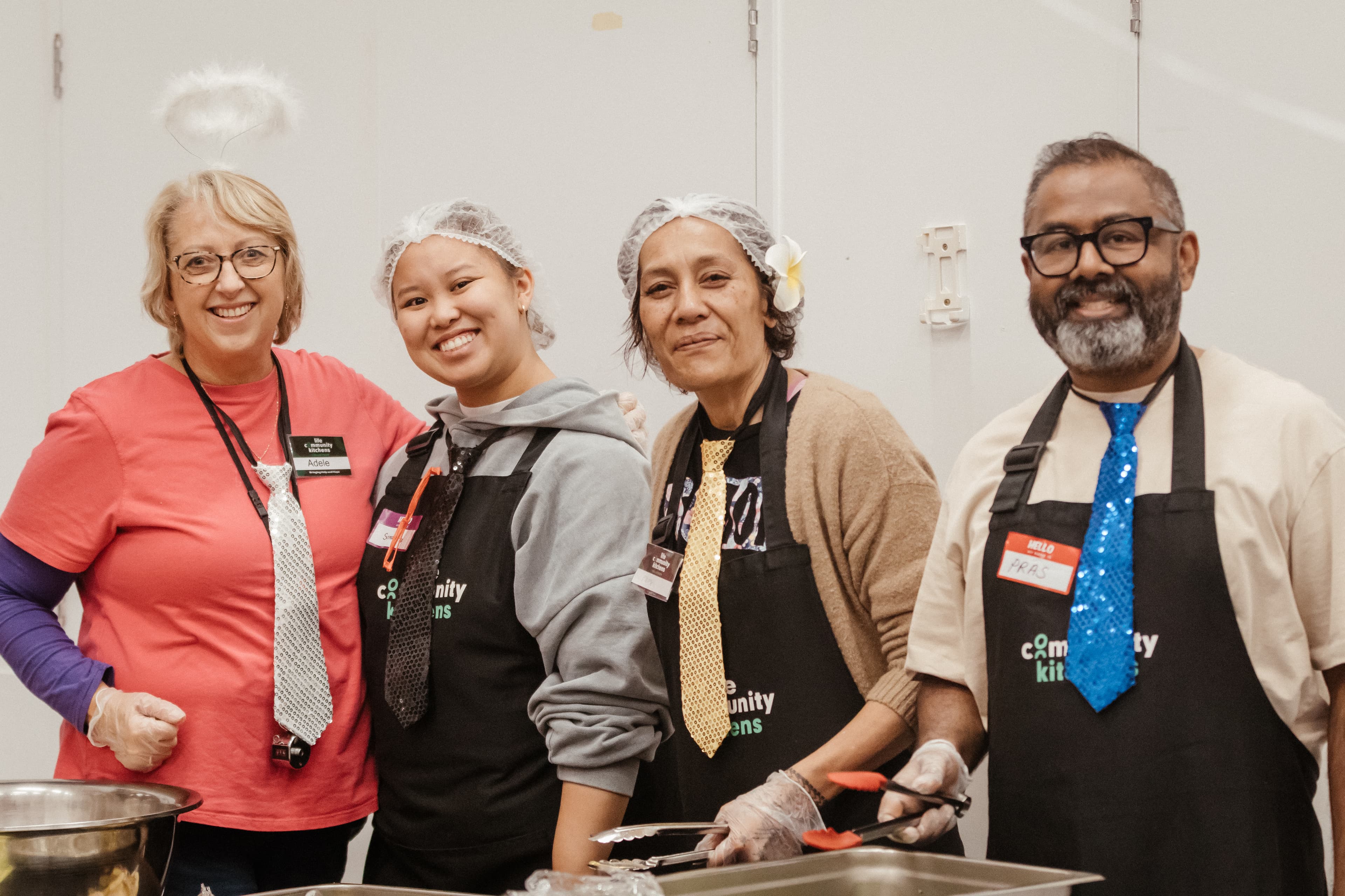 Four volunteers at LIFE Community Kitchens serving food and smiling at the camera with hair nets and dress up ties on
