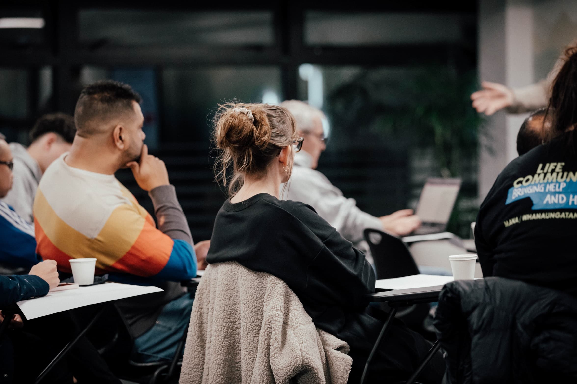 People sitting in a classroom at Night School with focus on a woman an two men sitting at desks with notebooks or laptops open