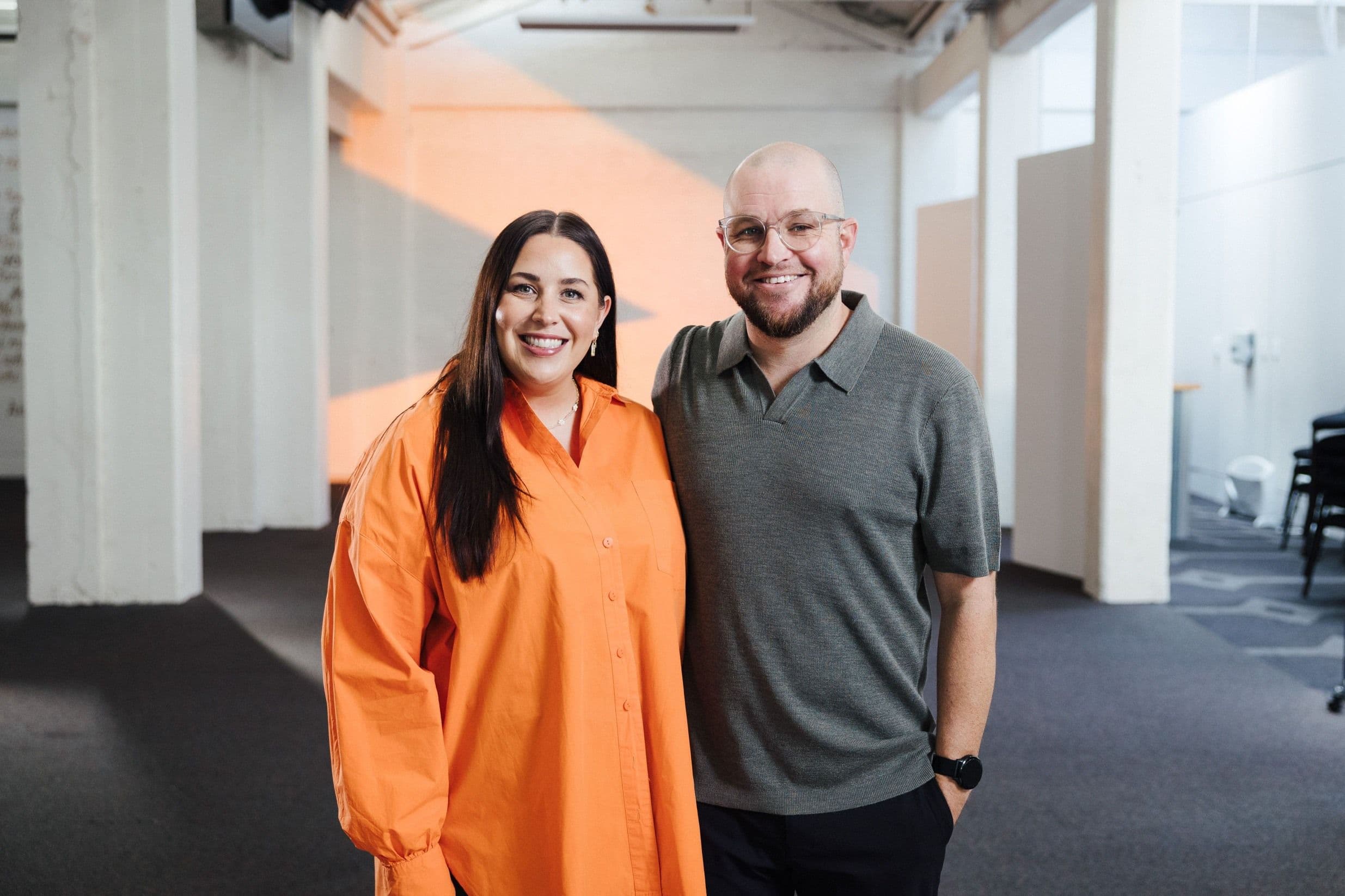 Ps Missy and Ps Luke standing close together and smiling at the camera with the orange chevron (>) projected in orange on a wall behind them
