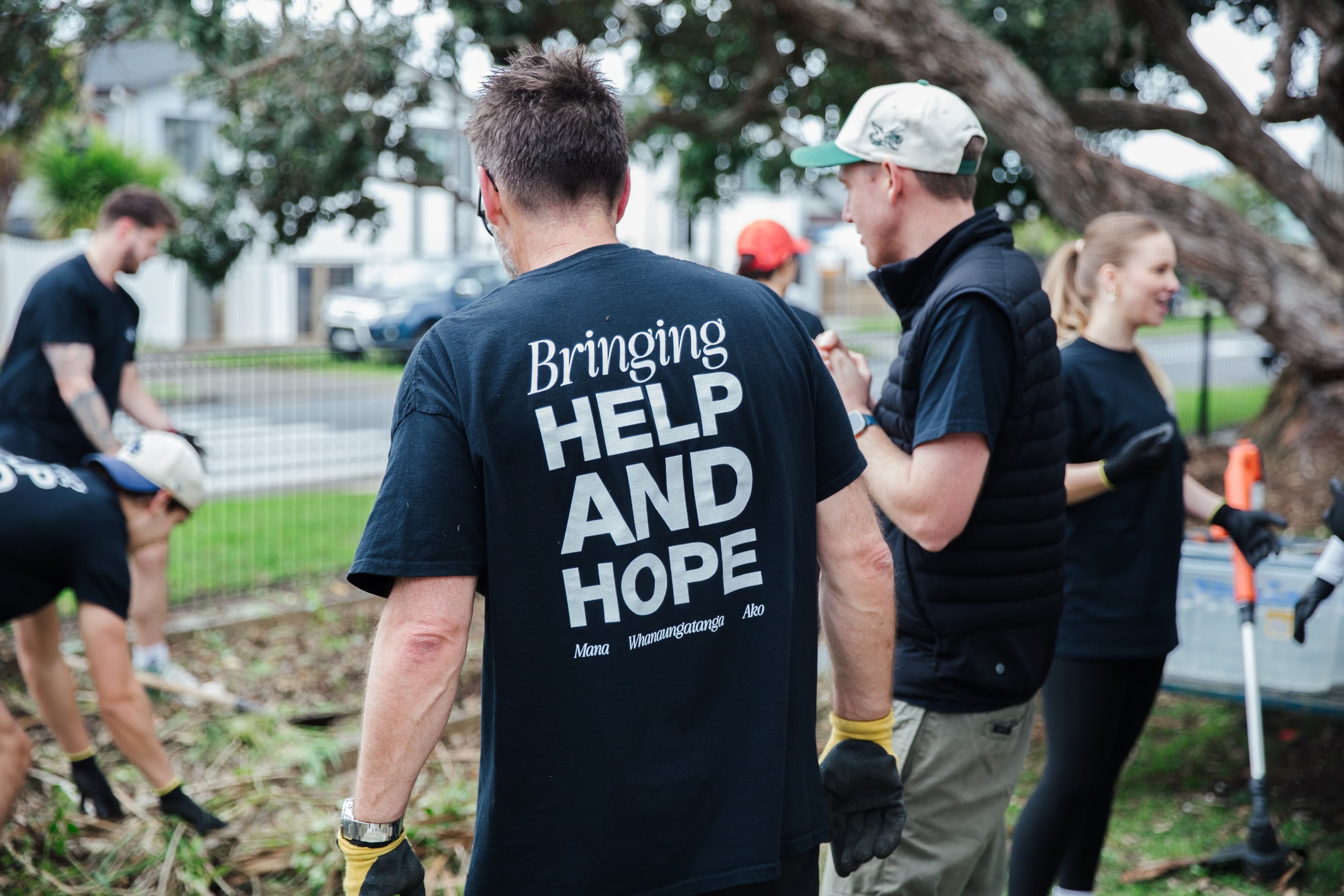 Two men facing away from the camera helping to work on a garden with black t-shirts saying Bringing Help and Hope to the Community for Help and Hope Week