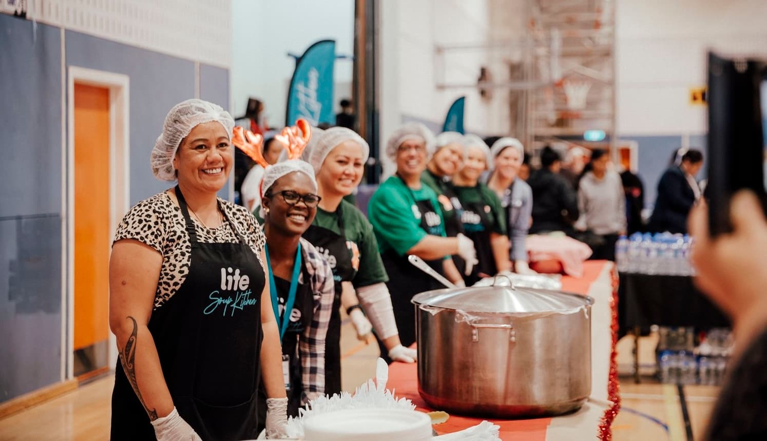 Volunteers standing behind tables of food prepared to serve diners at a LIFE Community Kitchen, all wearing hairnets and aprons